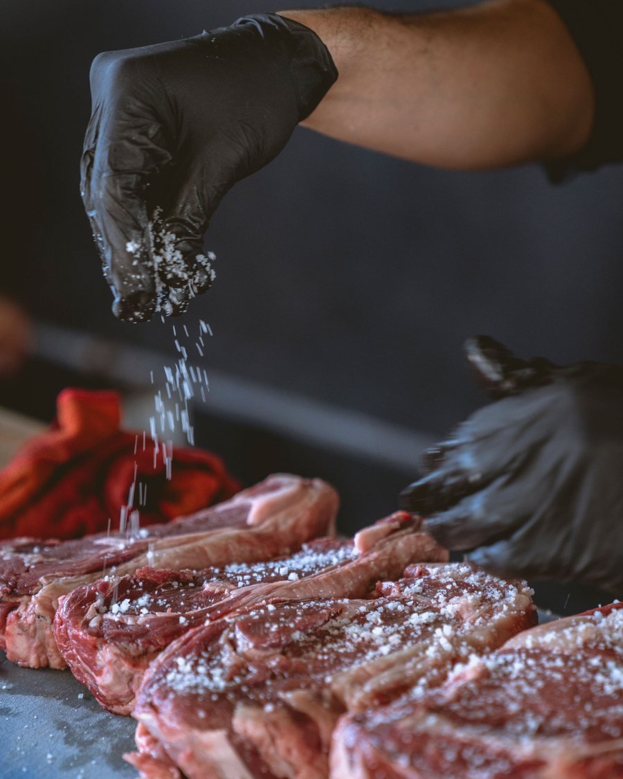 Main gantée de noir saupoudrant du gros sel sur des côtes de bœuf crues, en cuisine.